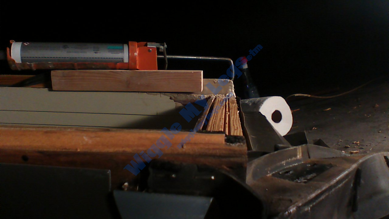 A view showing the gap still needed to fill to bring the missing window frame up high enough to seal against the glass.  The 1x2 wood piece is level with the good part of the frame being held by the weight of my caulking gun, revealing the gap between the bottom of the 1x2's right end and the window frame's new wood fill.